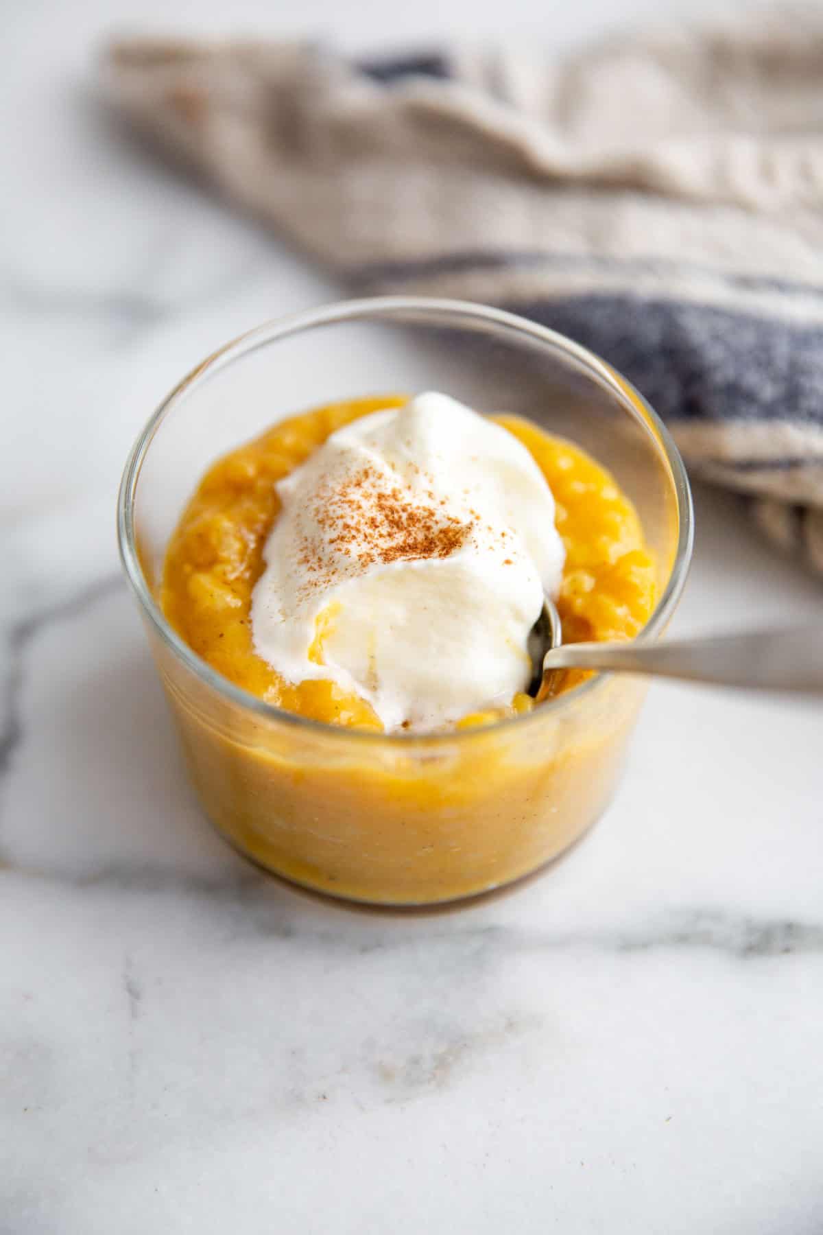 a spoon resting inside a jar of pumpkin pudding topped with whipped cream and pumpkin pie spice. 