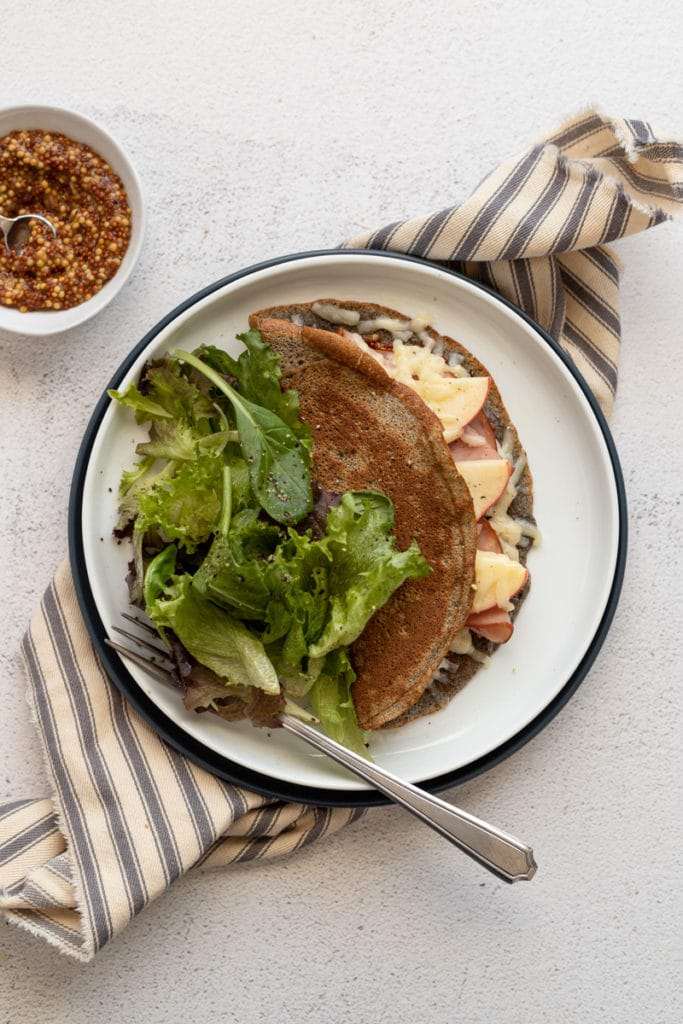 A crepe on a plate stuffed with ham, cheese and apples, with a salad alongside.