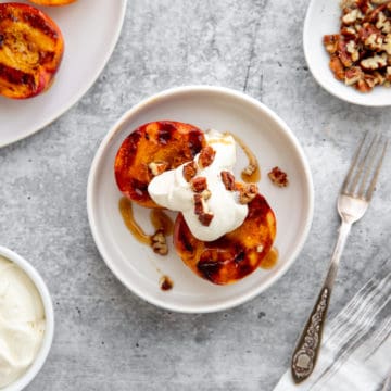 Grilled peaches on a plate topped with cardamom whipped cream and toasted nuts.