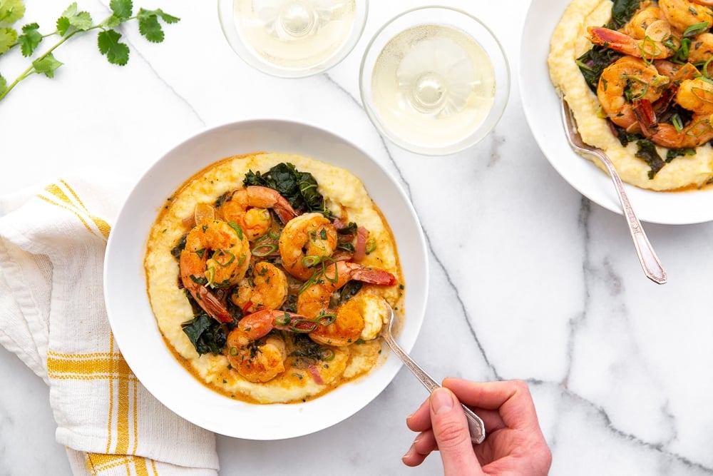 Coconut sriracha shrimp and kale over grits in serving bowls, with hand taking a bite with a fork