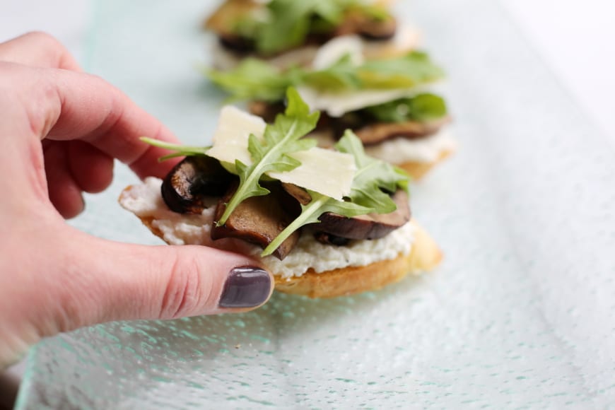 Hand grabbing a ricotta crostini topped with mushrooms and arugula.