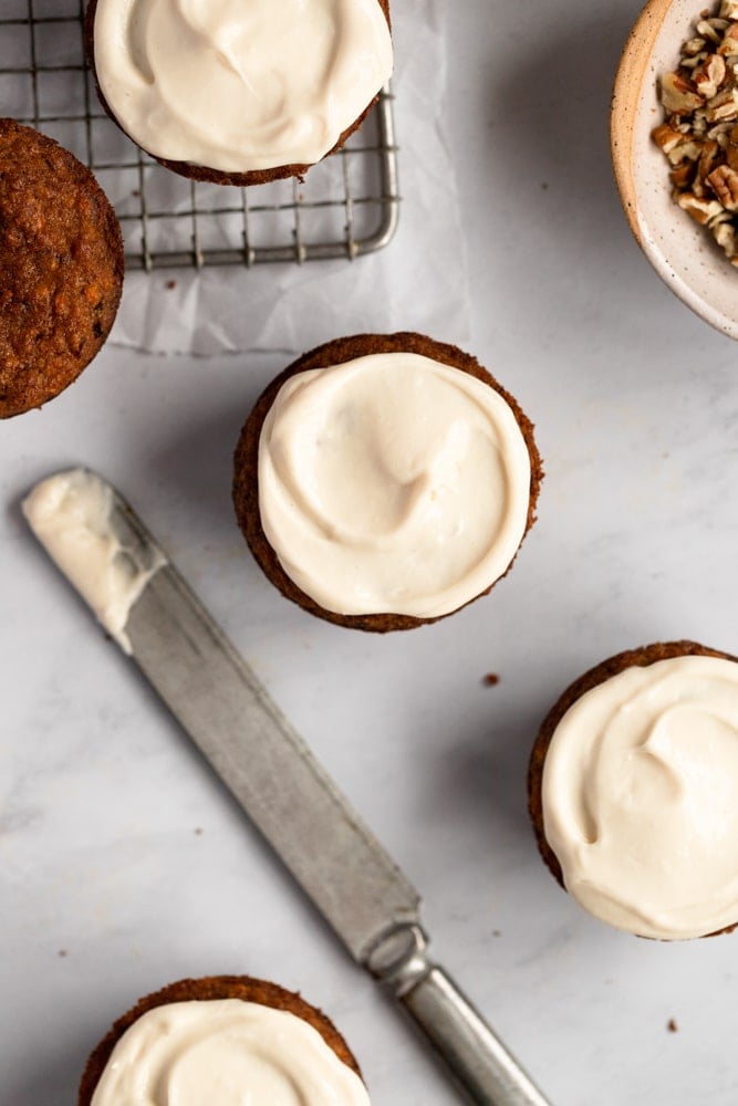 Healthy carrot cake muffins topped with maple cream cheese frosting, with a knife alongside.