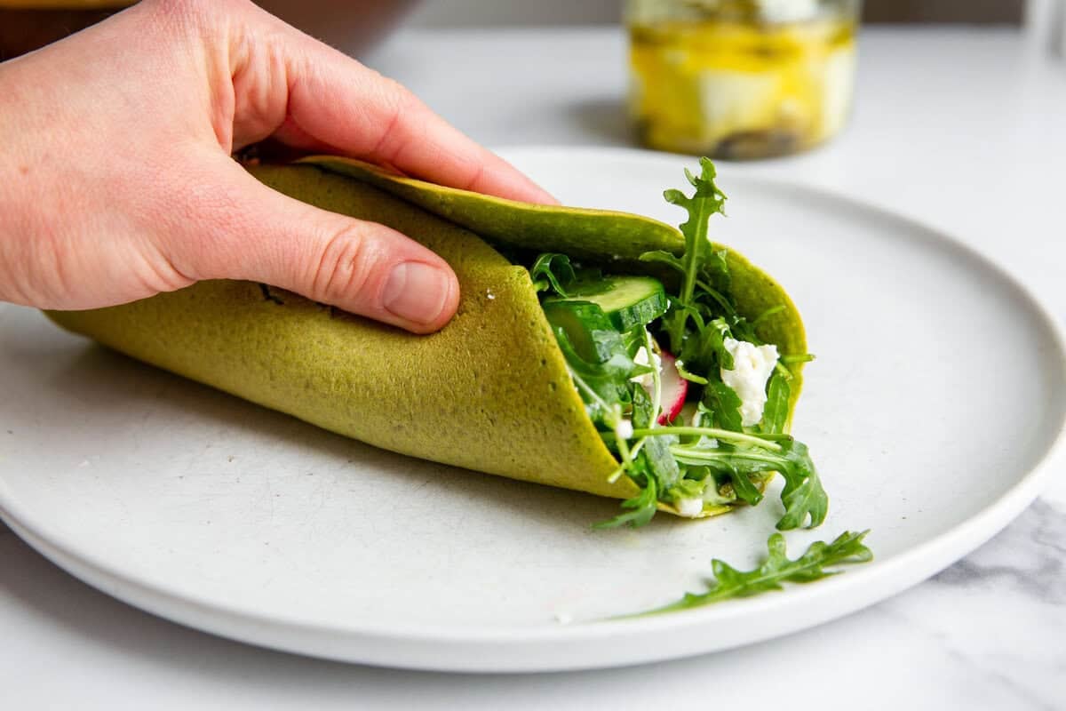 a hand holding a chickpea flatbread folded into a wrap stuffed with veggies and cheese. 