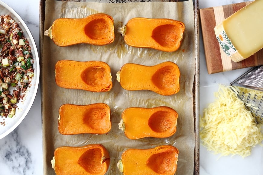 Process shot for the honeynut squash recipe showing roasted squash halves on a baking sheet with the quinoa stuffing and cheese alongside. 