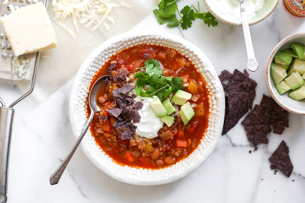 Overhead shot of a bowl of vegetable and quinoa chili on a table surrounded by toppings.