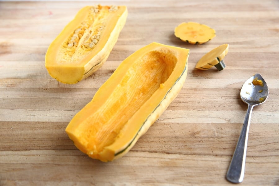 A halved delicata squash on a cutting board with a spoon for scraping out the seeds. 