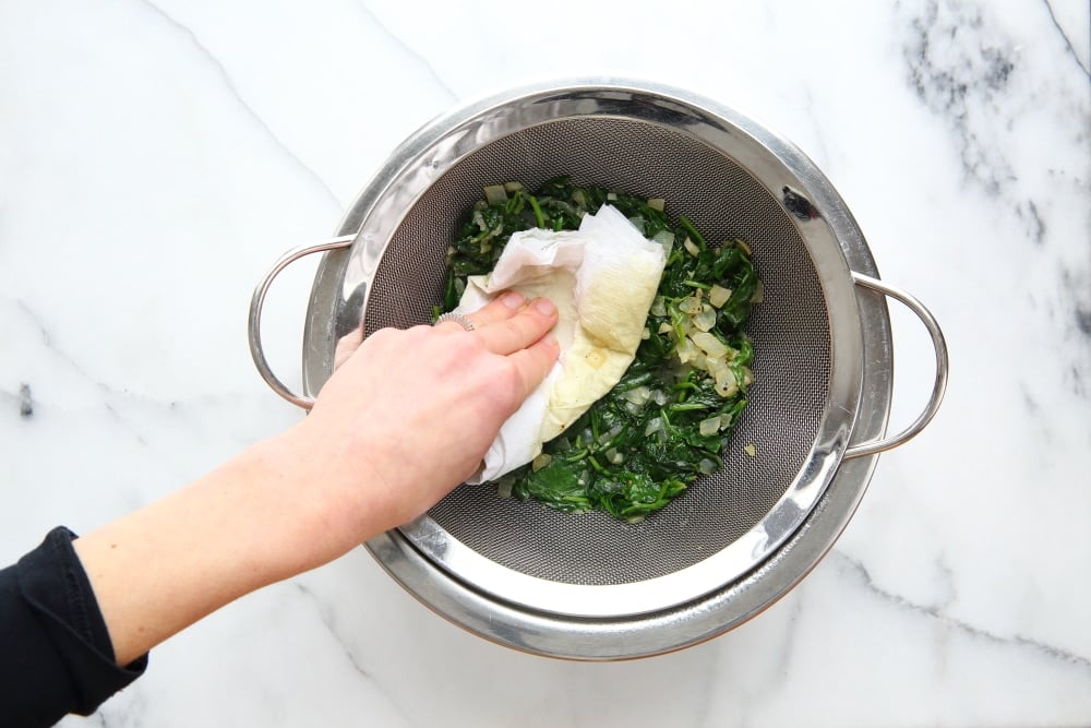 Process shot showing cooked spinach in a strainer, with a hand pressing out the liquid using a paper towel.