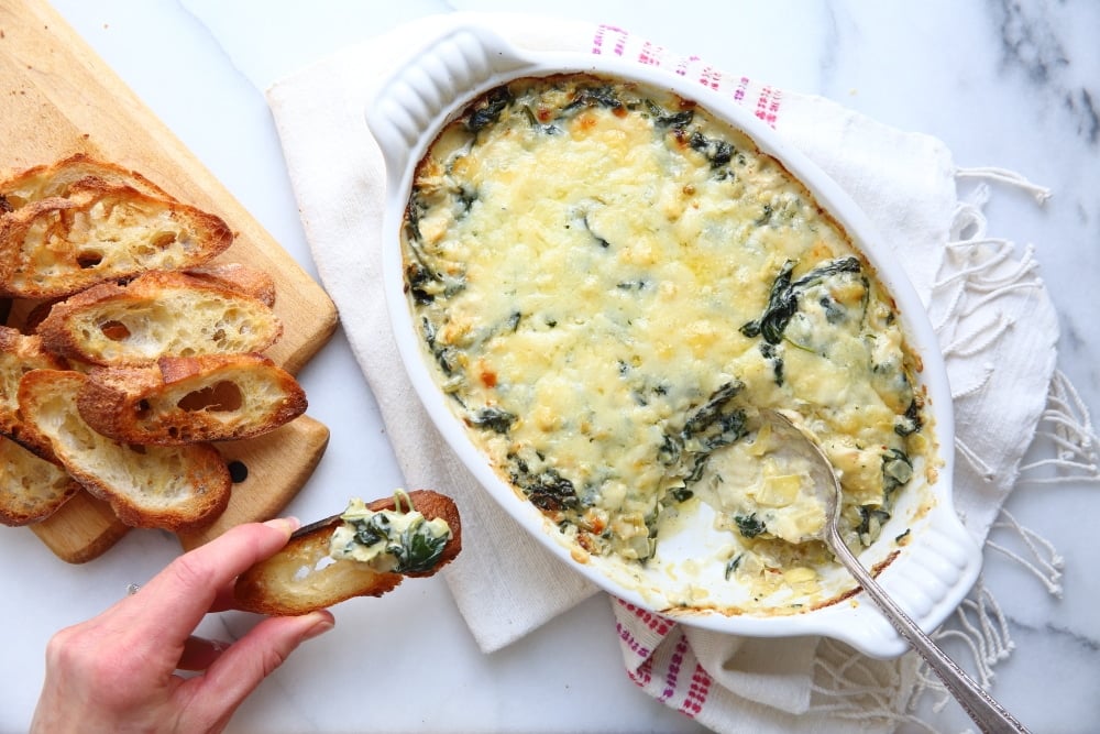 A hand holding a toasted crostini topped with warm spinach and artichoke dip.