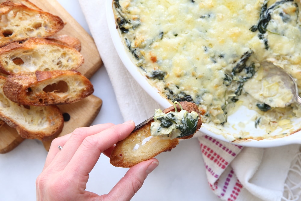 Close up of a hand holding a crostini topped with warm spinach and artichoke dip.
