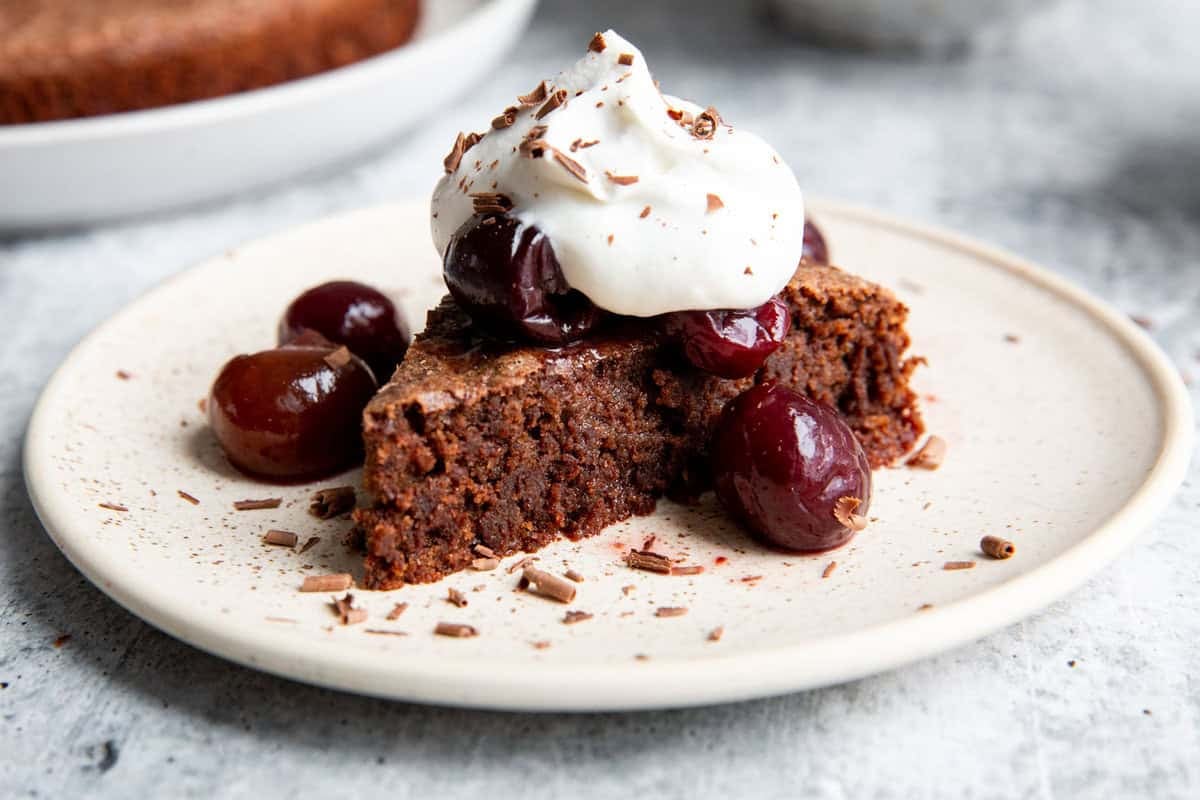 a slice of almond flour chocolate cake on a white plate topped with whipped cream and cherries