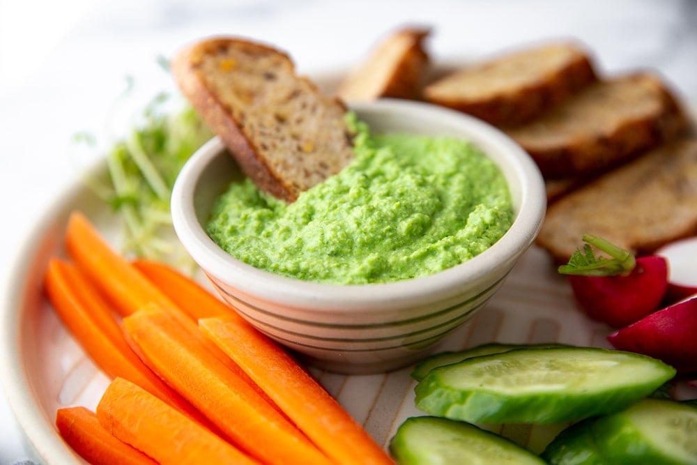 Pea dip in bowl on platter surrounded by vegetables and crostini for dipping