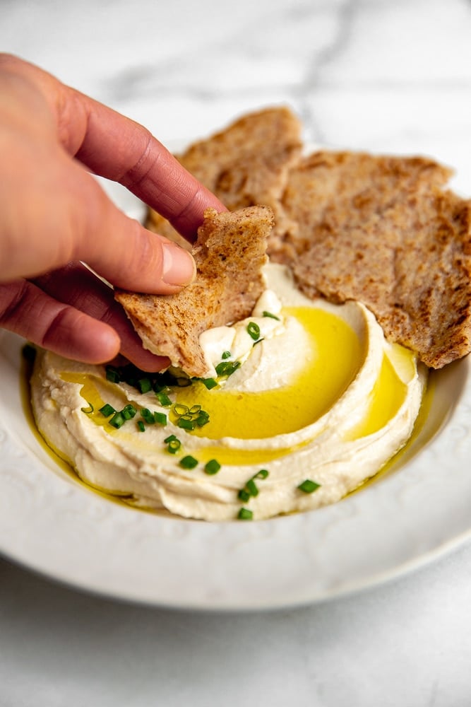 Hand dipping a cassava tortilla in a bowl of hummus 