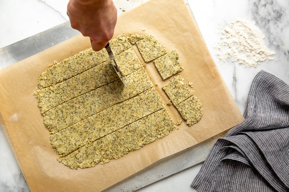 Seed cracker dough rolled out on a piece of parchment paper, with a hand cutting it into squares using a pizza cutter