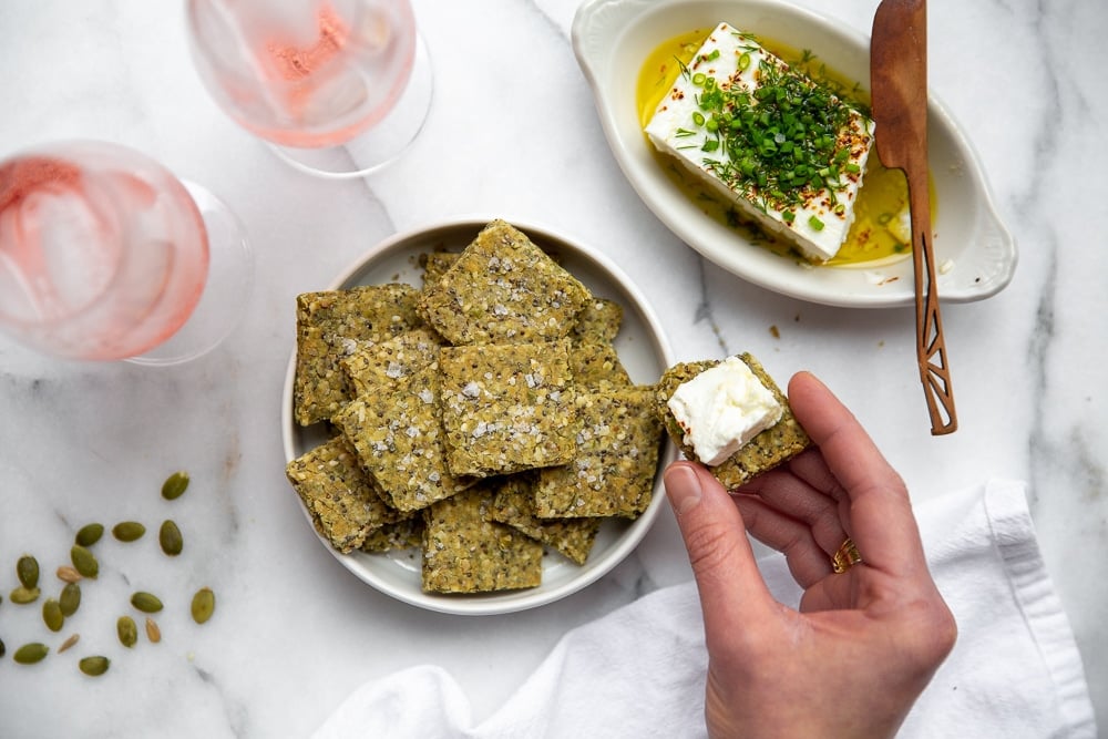 Hand holding a seed cracker topped with marinated feta 