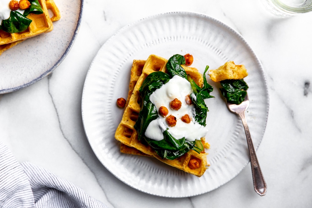 Two chickpea waffles stacked on plate, topped with sauteed spinach, yogurt and crispy chickpeas