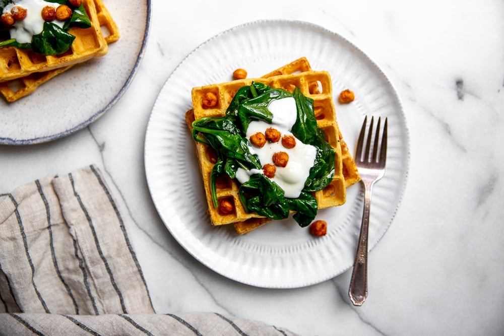 Two chickpea waffles stacked on plate, topped with sauteed spinach, yogurt and crispy chickpeas