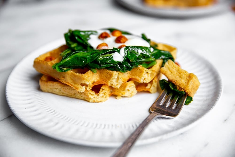 Chickpea waffles on plate with sautéed spinach, yogurt and crispy chickpeas, with fork taking a bite