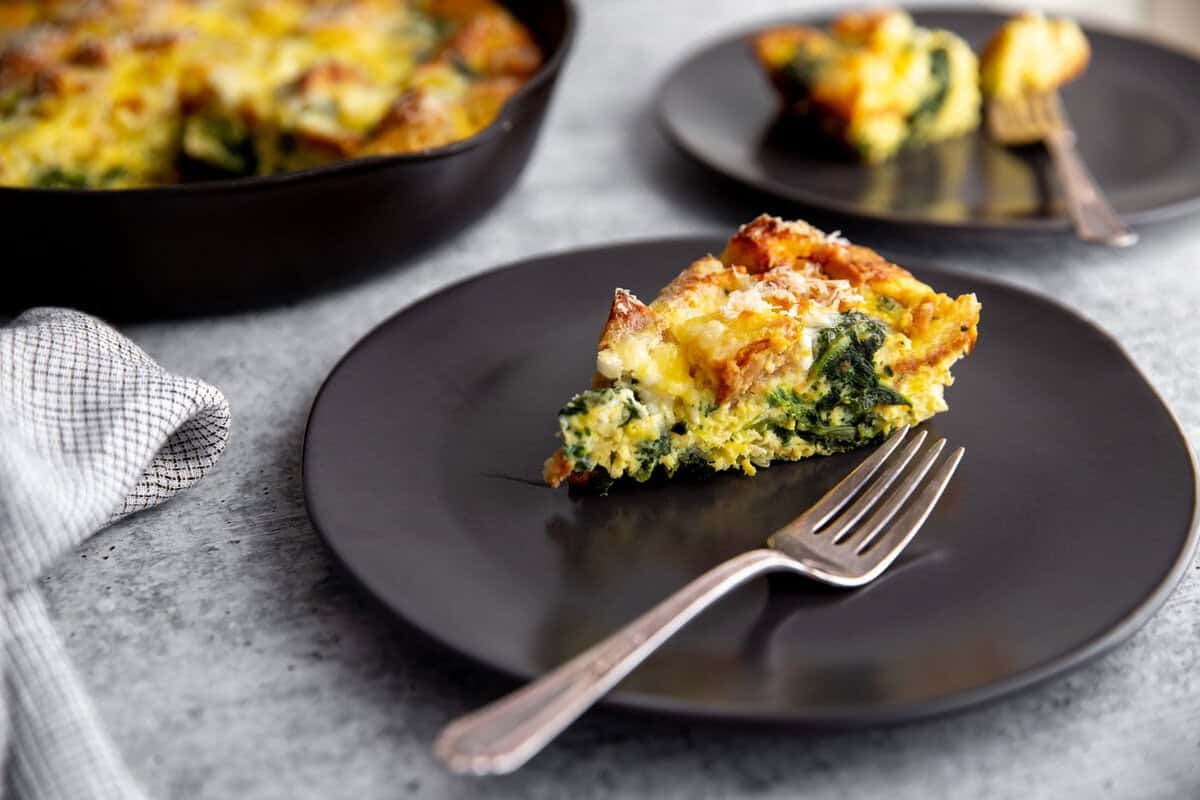 A slice of vegetable strata is served on a black plate with a fork, next to a plaid napkin. Another plate with a slice and a skillet with the remaining frittata are visible in the background on a gray surface.