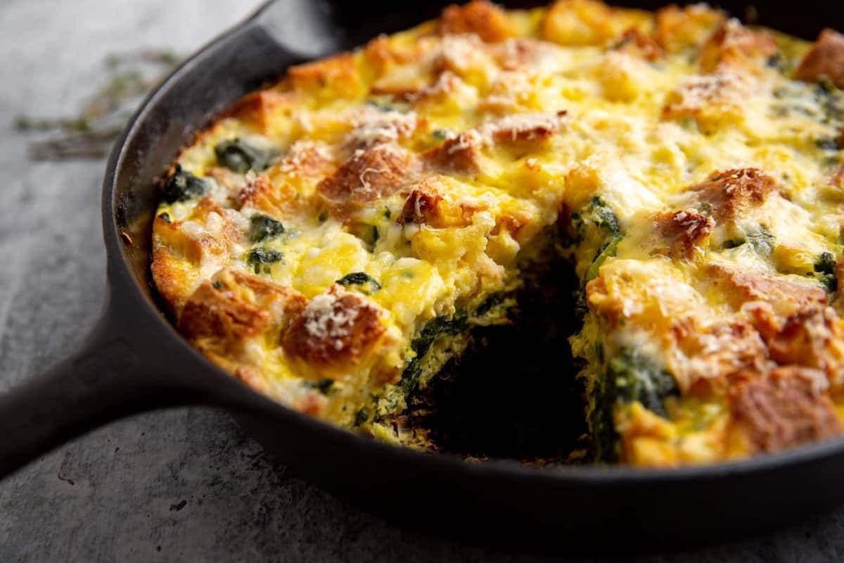 A close-up of a baked egg and spinach stratta in a black skillet. The dish features a golden-brown crust with visible pieces of bread and melted cheese on top. A slice has been removed, revealing layers of spinach and egg inside.