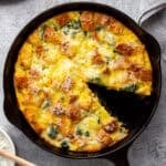 A cast iron skillet containing a baked vegetable strata with a golden-brown top. The strata is filled with spinach and pieces of bread, topped with melted cheese. A slice has been removed, revealing the texture inside. A small checkered cloth is nearby.