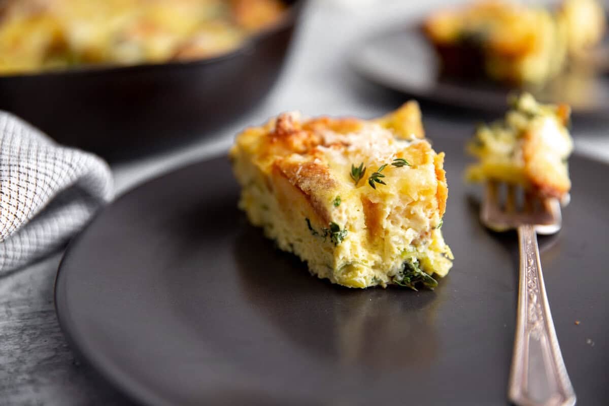 A slice of vegetable strata with herbs on a black plate, next to a fork. The dish includes visible cheese and greens, with another plate in the blurred background. A cloth napkin is partially visible on the left.