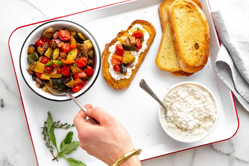 A tray holding a bowl of ratatouille, cottage cheese and toasted slices of bread, with a hand topping one of the pieces of bread