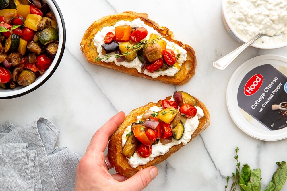 Hand grabbing a toast topped with cottage cheese and ratatouille, with bowls of ratatouille and cottage cheese on the side