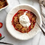 A serving of gluten free strawberry crisp in a serving bowl with a dollop of whipped cream