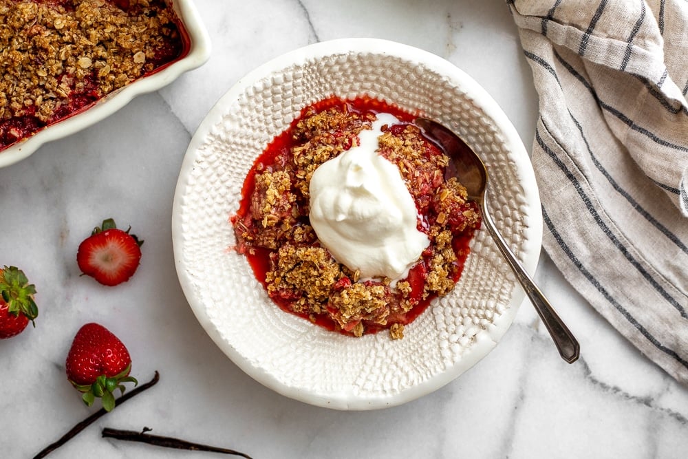 A serving of gluten free strawberry crisp in a serving bowl with a dollop of whipped cream