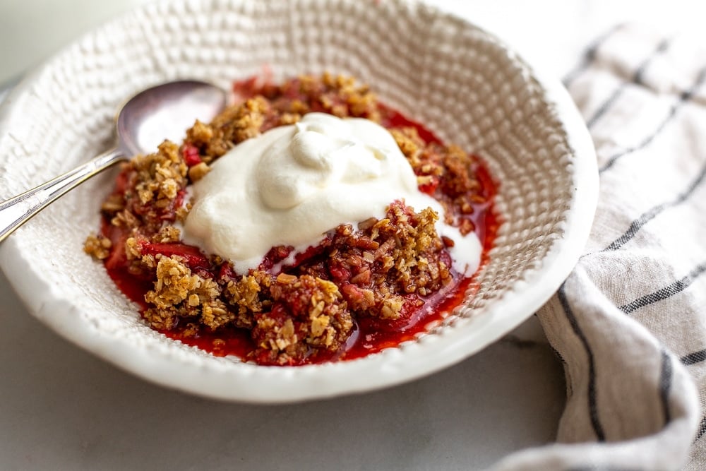 Strawberry crisp in bowl with whipped cream on top and serving spoon alongside