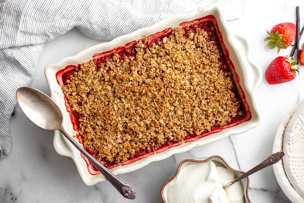 Gluten free strawberry crisp in baking dish with serving spoon and a bowl of whipped cream alongside