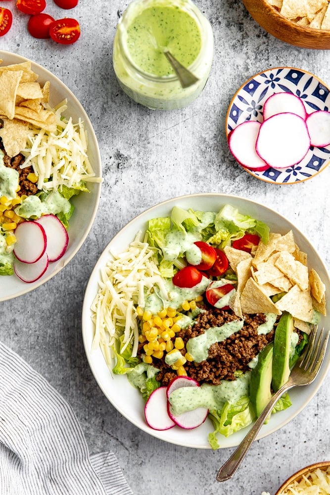 Healthy taco salad in bowl, with toppings alongside, including shredded cheddar cheese, radishes, tomatoes, tortilla chips and creamy cilantro lime dressing