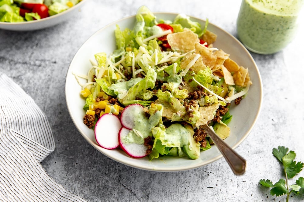 Healthy taco salad tossed in bowl with fork, with creamy cilantro lime dressing on the side