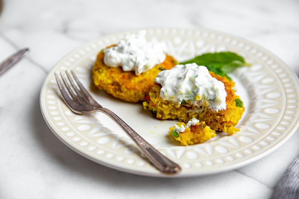 Two millet cakes on serving plate, topped with cucumber mint raita, with a fork alongside and a bite taken out