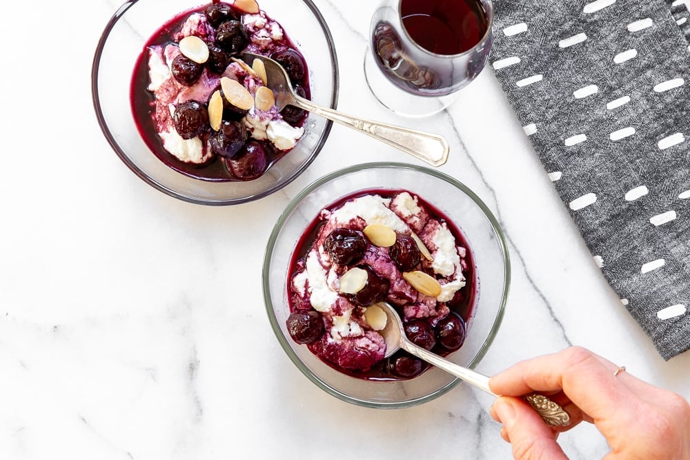 Two bowls of cherries in port over fresh ricotta, with a hand holding a spoon. 