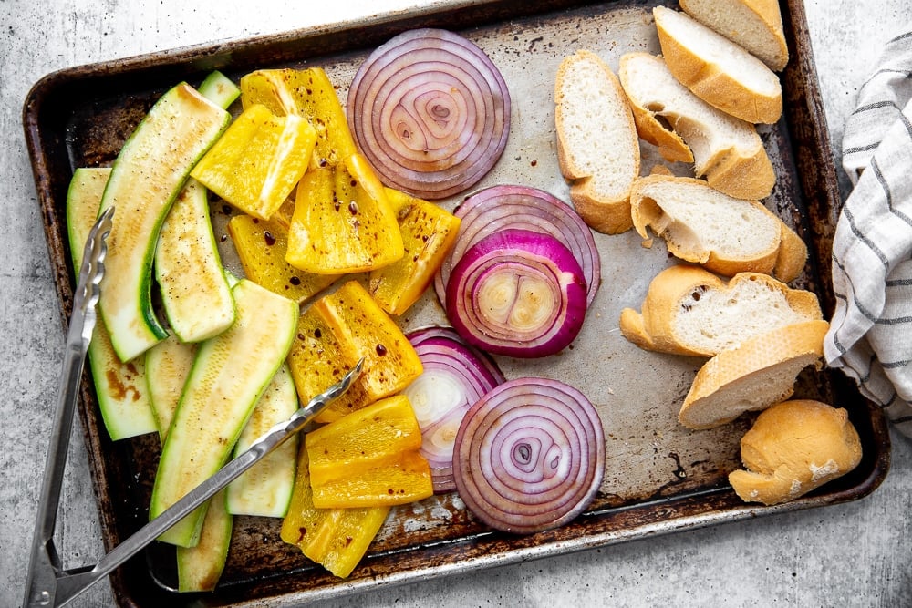 Process shot showing vegetables and bread spread out on a sheet pan, ready to go on the grill. 