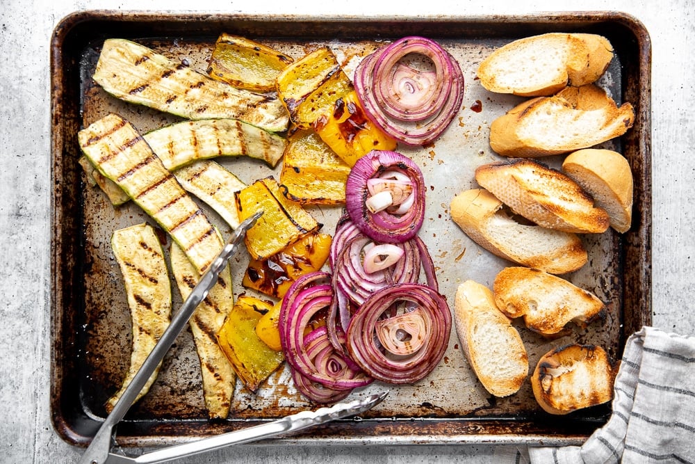 Process shot showing grilled vegetables and bread spread out on a sheet pan.