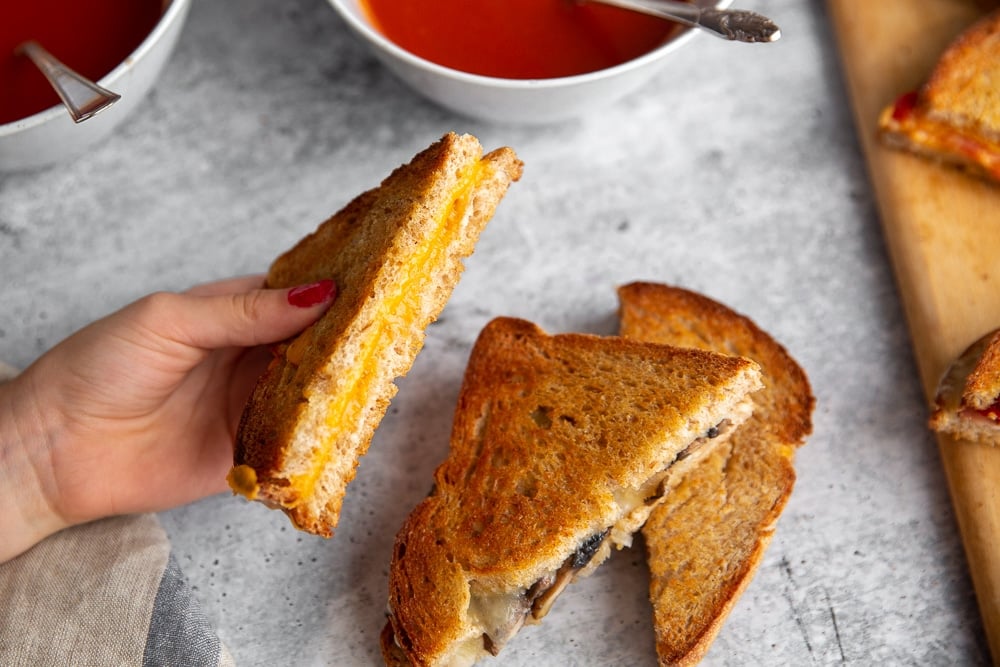 Hand holding a grilled cheese half, with a bowl of soup in the background.