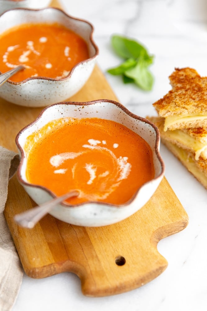 Two bowls of tomato soup on a wooden serving bowl, with a grilled cheese sandwich alongside.