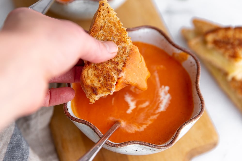 Hand dipping a grilled cheese sandwich into a bowl of tomato soup.