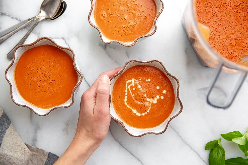 Overhead shot of a hand grabbing a bowl of tomato soup, with a Vitamix container alongside. 