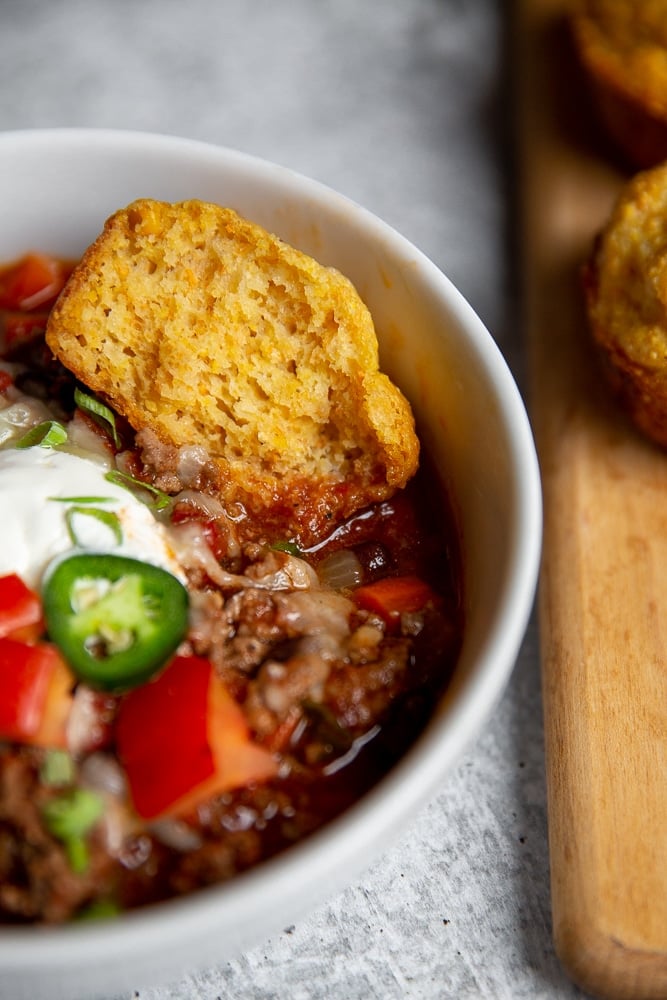 Close-up of a cornbread muffin dunking into a bowl of chili.