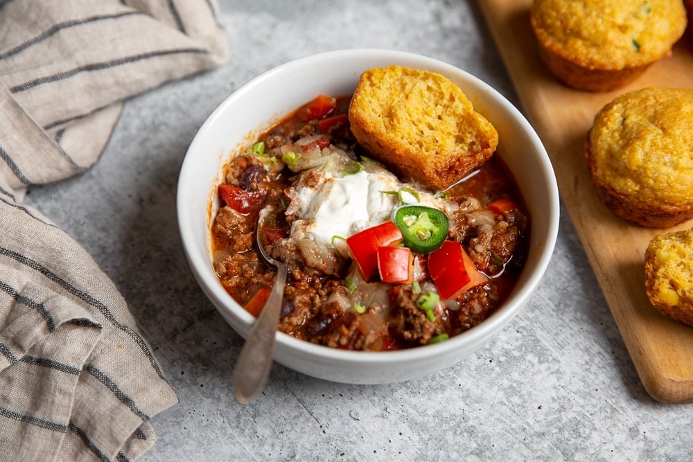 Bowl of chili with jalapeno cheddar cornbread muffins alongside.