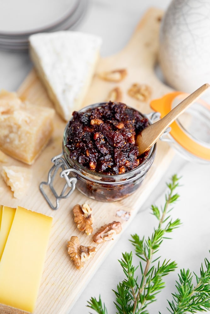 Fig chutney in a jar on a serving board with cheeses alongside. 