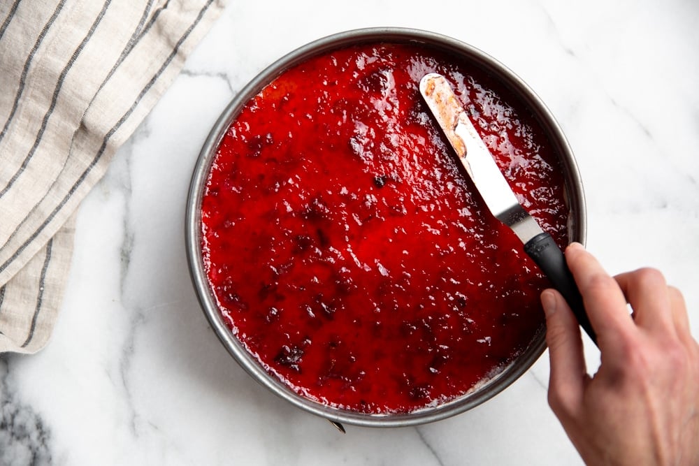 Hand using an off-set spatula to spread cherry jam over the cheesecake. 