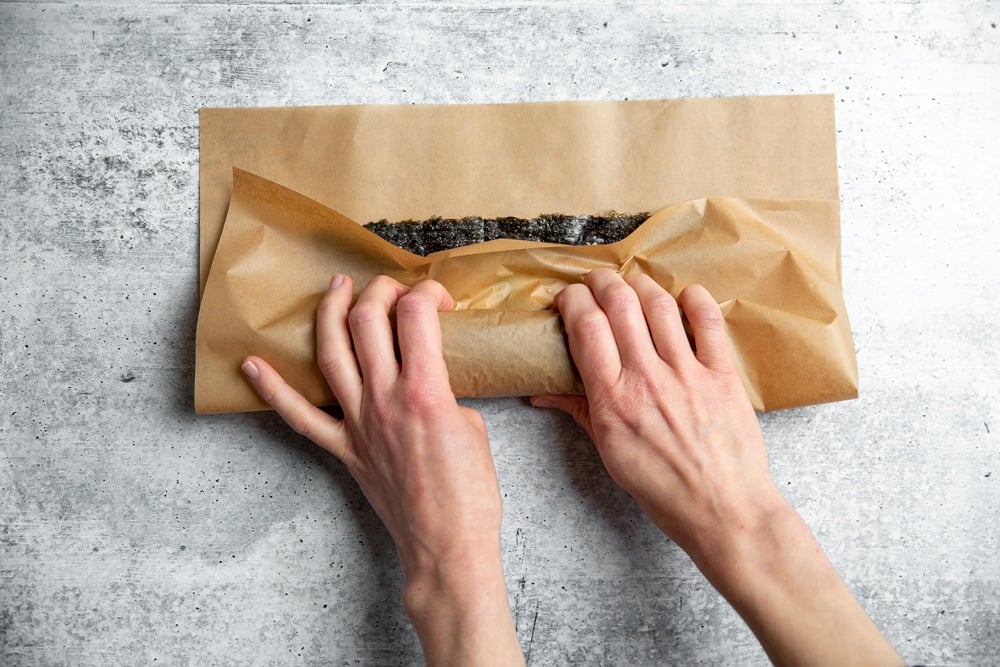 Hands using a piece of parchment paper to roll a salmon sushi roll. 