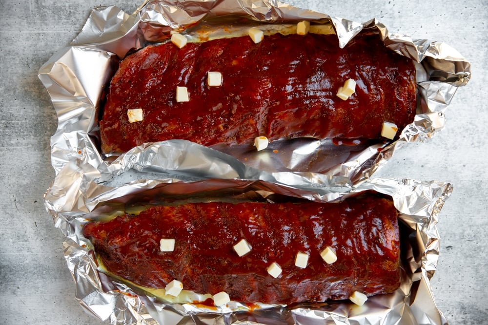Process shot showing smoked ribs in foil packets dotted with butter.