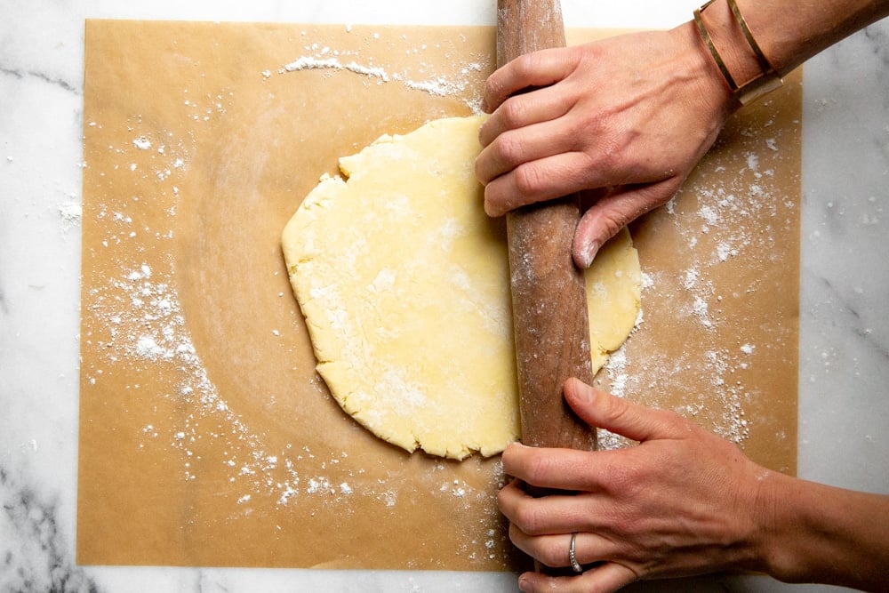 Process shot showing hands rolling out a piece of pie dough. 