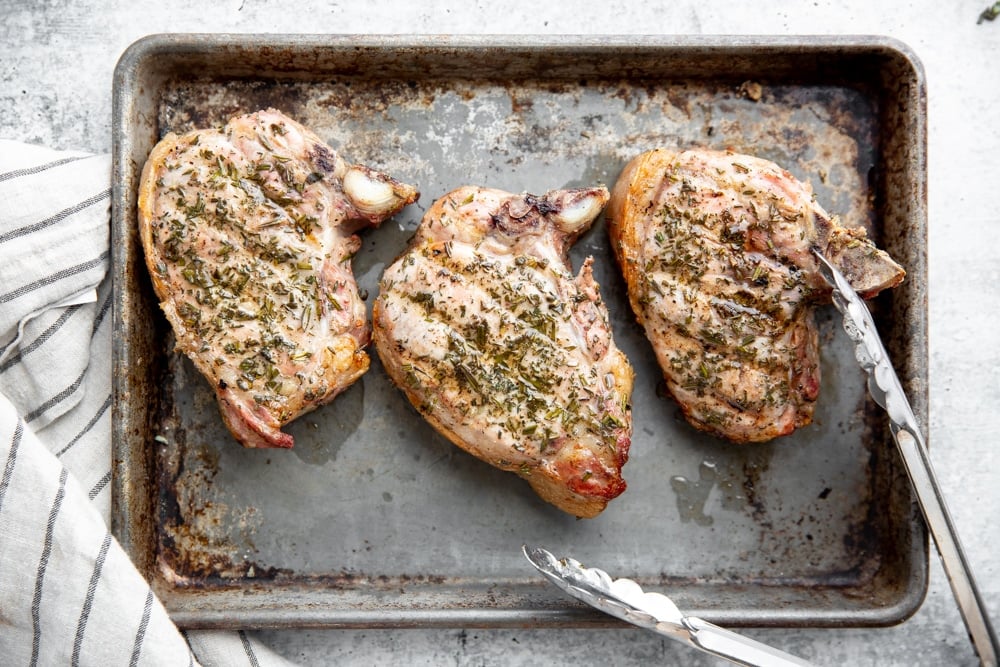 Grilled thick cut pork chops on a sheet tray with tongs. 