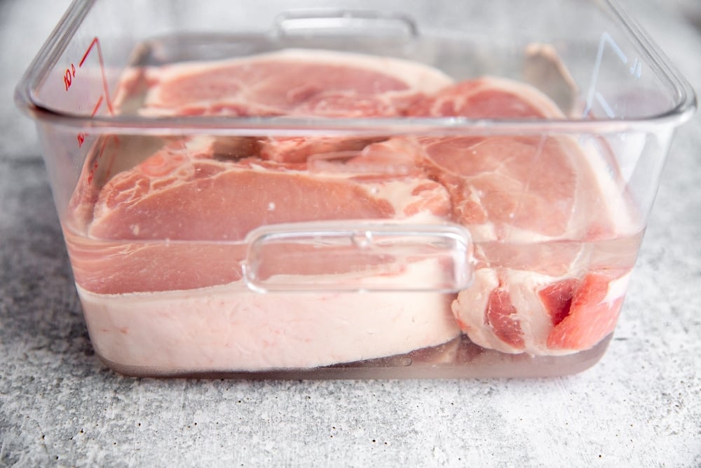 Process shot showing thick cut pork chops brining in a clear container. 